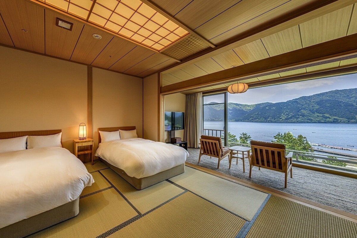 A hotel room with two beds, a TV, a balcony with a view of a lake and mountains, and traditional Japanese tatami flooring.