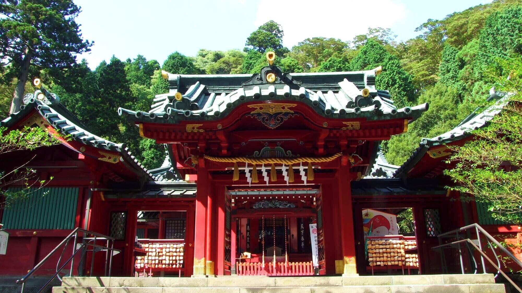 A traditional Japanese temple with a red entrance gate, a staircase leading up to it, and a pagoda in the background.