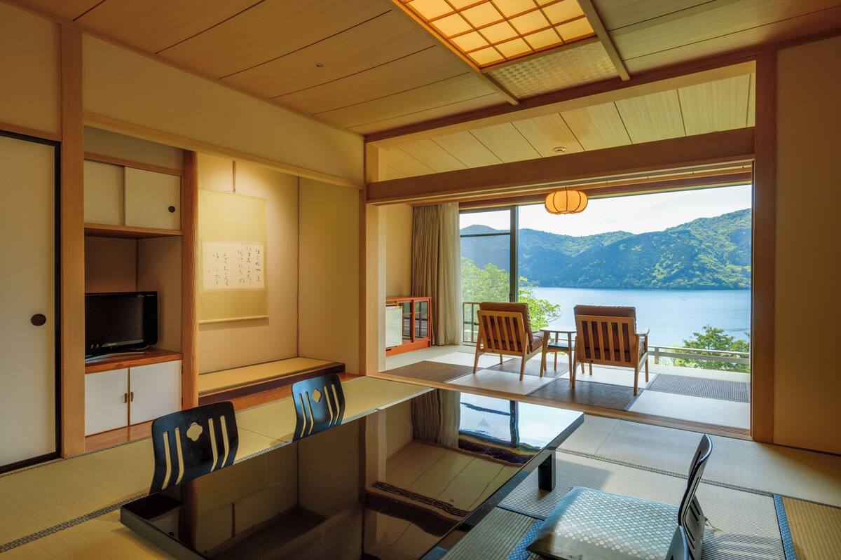 A traditional Japanese room with tatami flooring, a low table, chairs, a television, and a view of a lake and mountains.