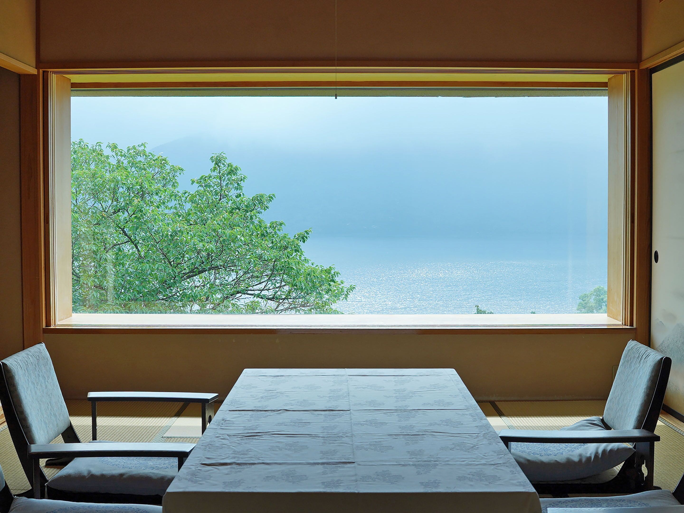 A room with a large window overlooking a tree and a body of water. There is a table with chairs in front of the window.
