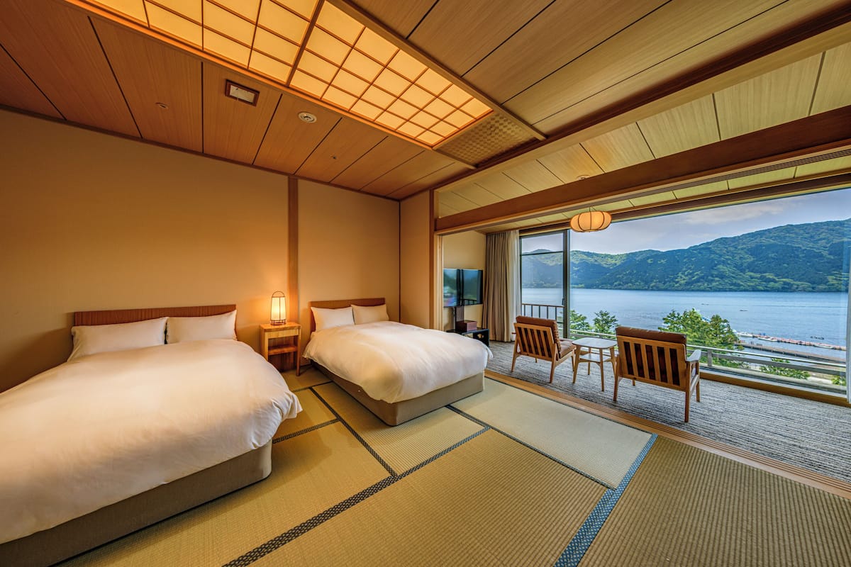 A hotel room with two beds, a sitting area with a view of a lake and mountains, and traditional wooden architecture.