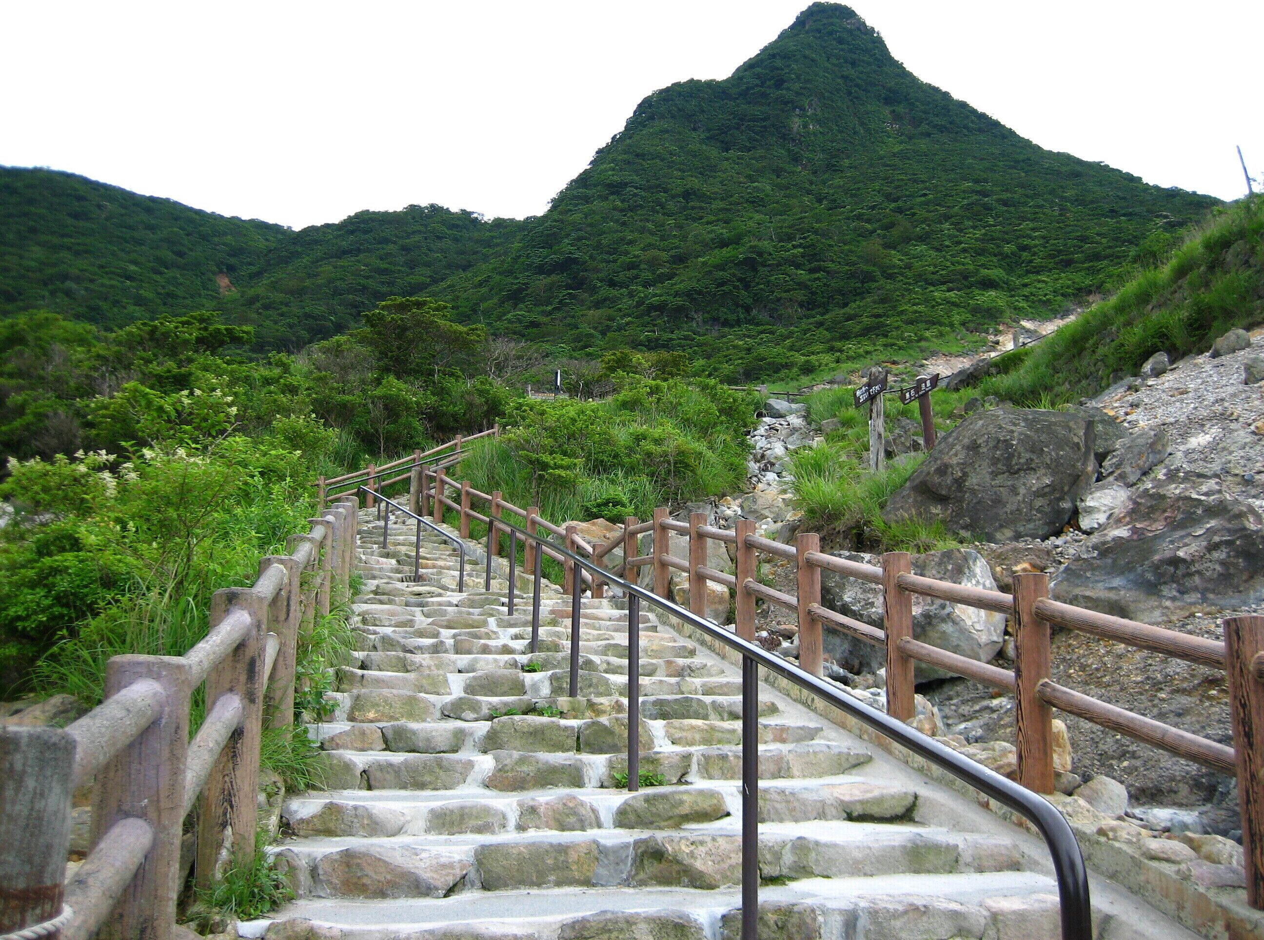 A stone staircase with wooden railings leading up a mountain path, surrounded by lush greenery.