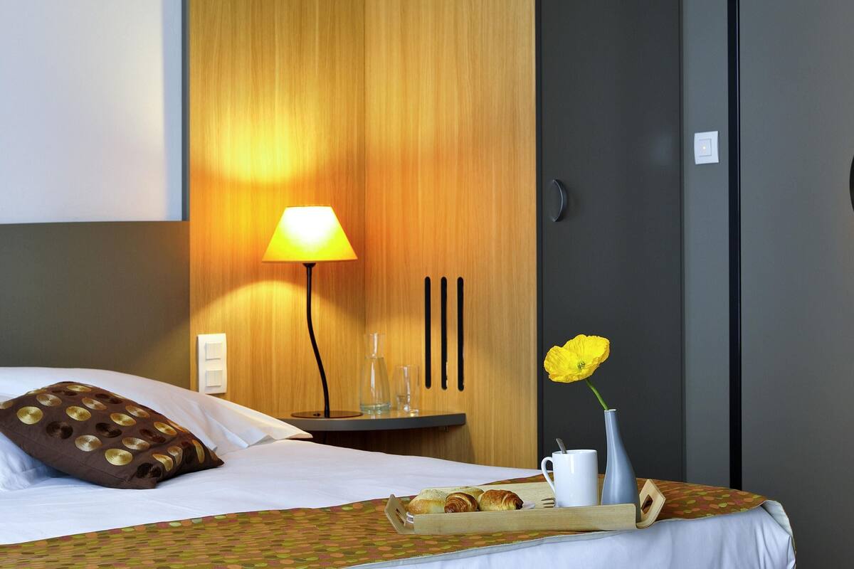 Een hotelkamer met een bed, een nachtkastje met een lamp, een vaas met een bloem en een dienblad met gebak.