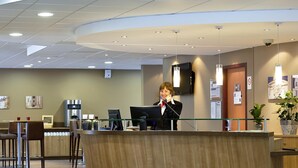 A reception desk with a woman standing behind it, a flat-screen TV mounted on the wall, and a potted plant to the right.