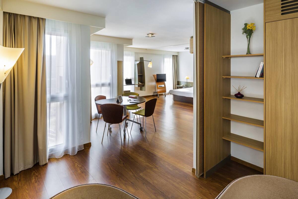 Een moderne hotelkamer met een houten vloer, een zithoek met een tafel en stoelen, een bed, een televisie en een boekenkast.