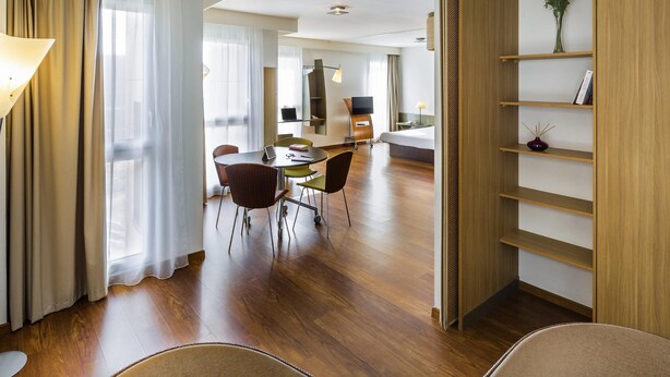 A modern hotel room with a wooden floor, a dining area with a table and chairs, a bed, a TV, and a bookshelf.
