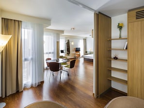 Una habitación de hotel moderna con suelo de madera, una zona de comedor con mesa y sillas, cama, televisor y estantería.