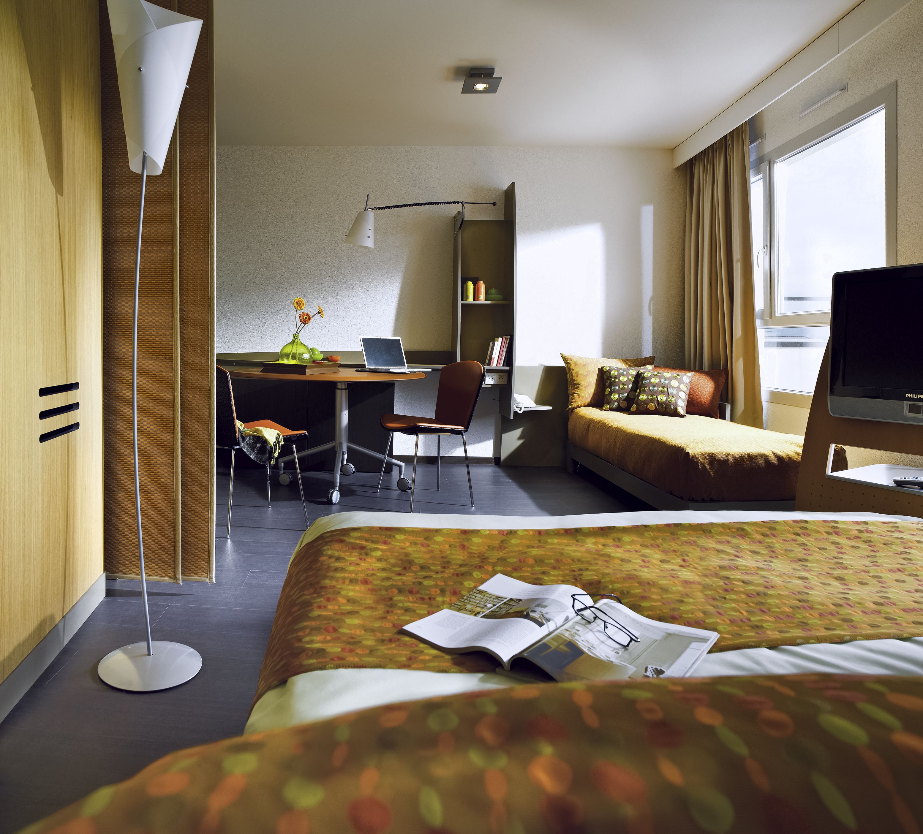 Une chambre d’hôtel avec un lit, un bureau avec un ordinateur portable, une chaise, une télévision et une fenêtre avec des rideaux.