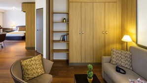 A modern hotel room with a wooden wardrobe, a sofa, a chair, a small table with a plant, and a lamp.