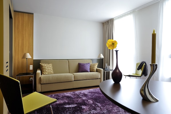 Una habitación de hotel moderna con un sofá, una mesa de comedor, una alfombra púrpura y un florero con una flor amarilla.