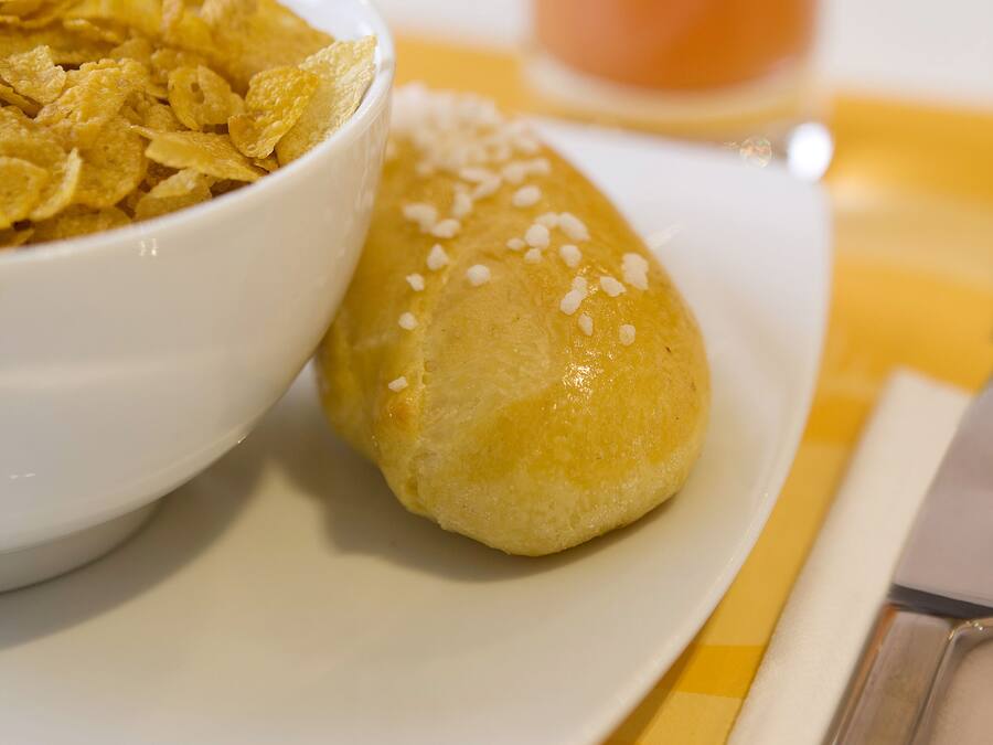 Un tazón de cereales y un panecillo sobre un plato, con un vaso de jugo al fondo.