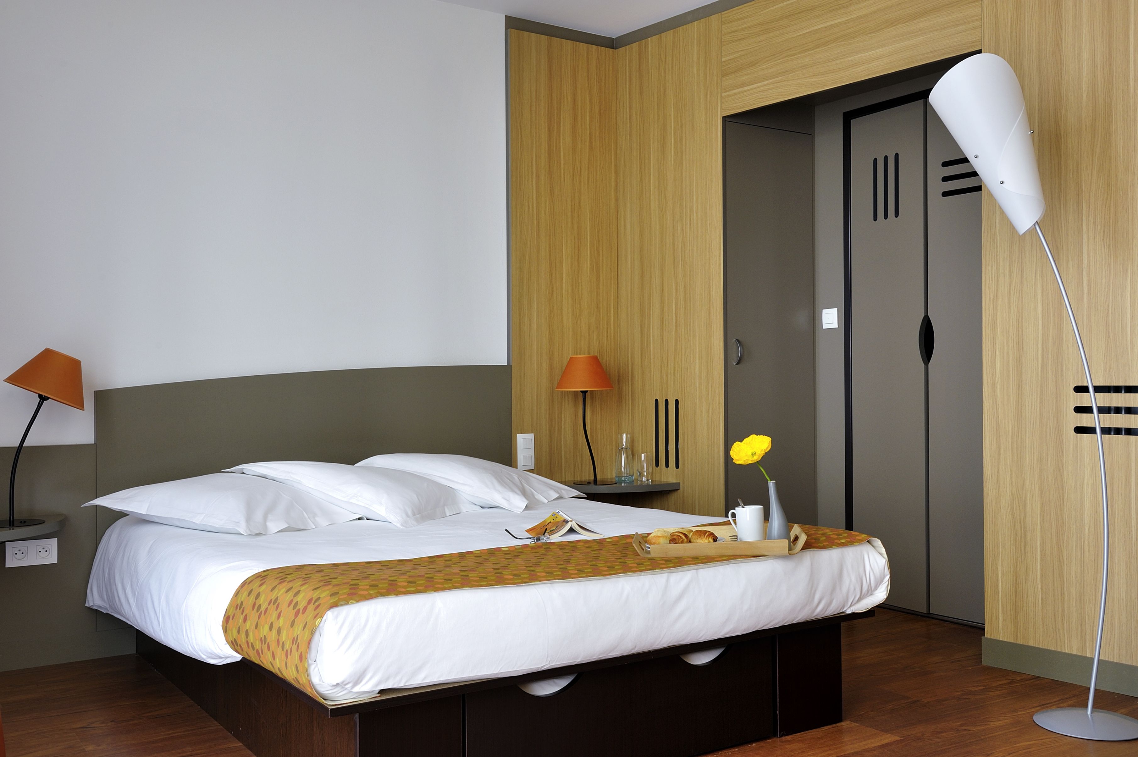 A neatly made bed with white linens, a bedside table with a lamp, and a wooden wardrobe.
