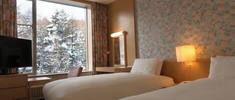 A hotel room with two beds, a TV, a desk, and a window with a view of snow-covered trees.