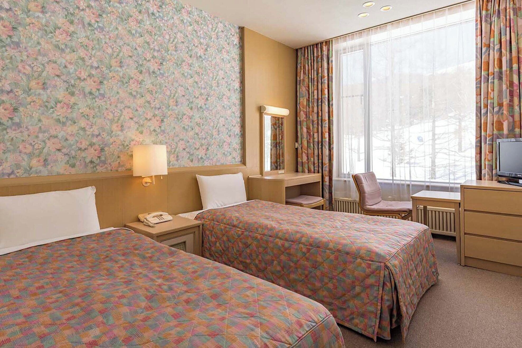 Habitación de hotel con dos camas, un escritorio, una silla, un televisor y un ventanal con cortinas.