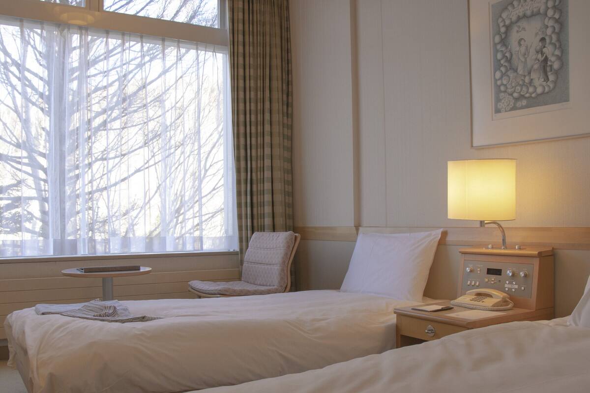 Habitación de hotel con dos camas, una mesita de noche con lámpara, una silla y un ventanal con cortinas translúcidas.