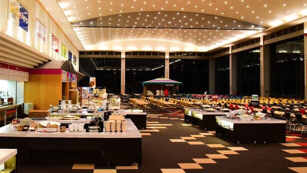 A large conference hall with multiple tables and chairs, a high ceiling with numerous lights, and a stage area.