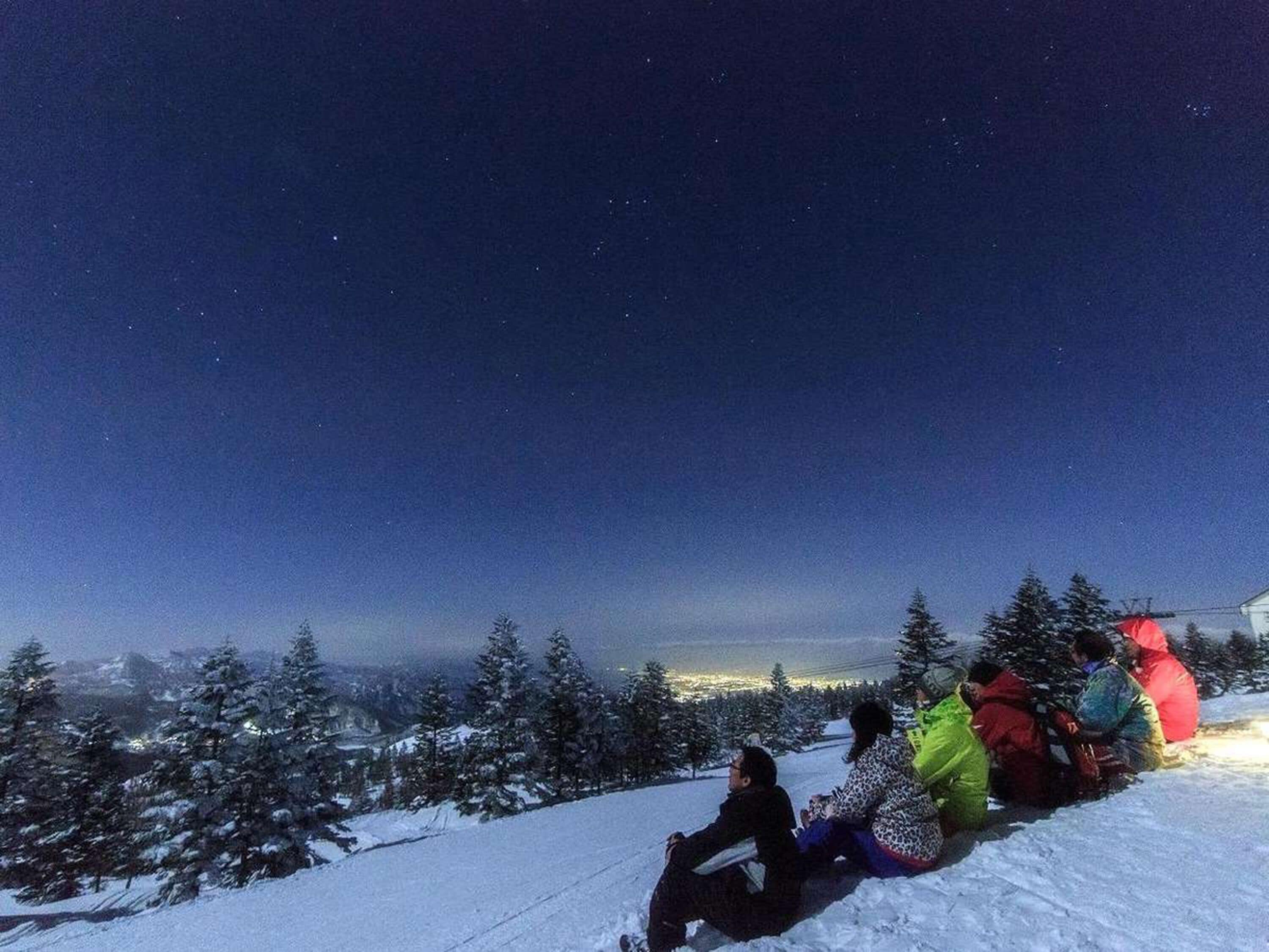 A group of people sitting on a snowy slope at night, overlooking a cityscape.