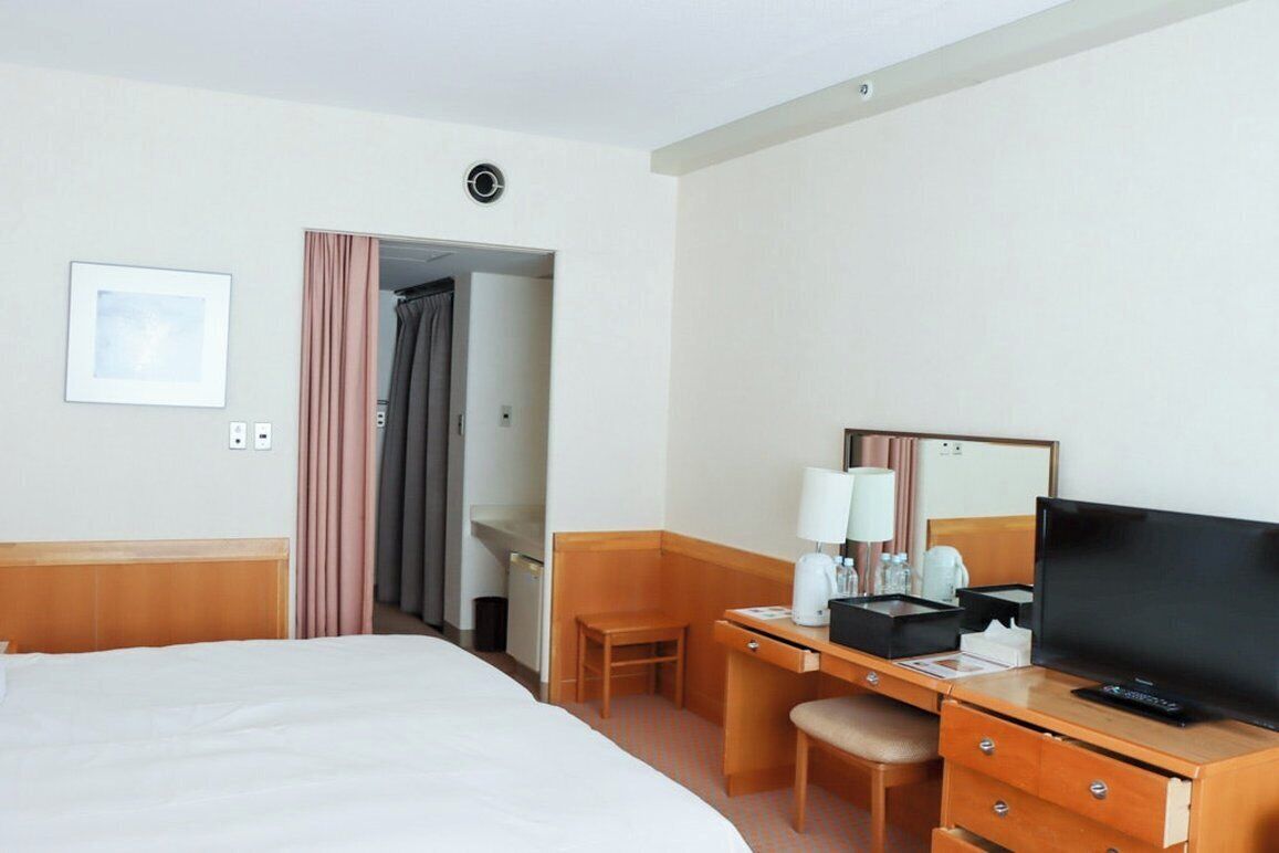 A hotel room with a bed, a desk with a television, a chair, a desk lamp, and a small table with a cup and a remote control.