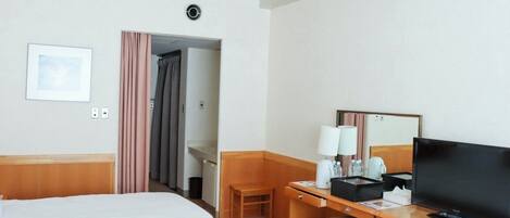 A hotel room with a bed, a desk with a television, a chair, a desk lamp, and a small table with a cup and a remote control.