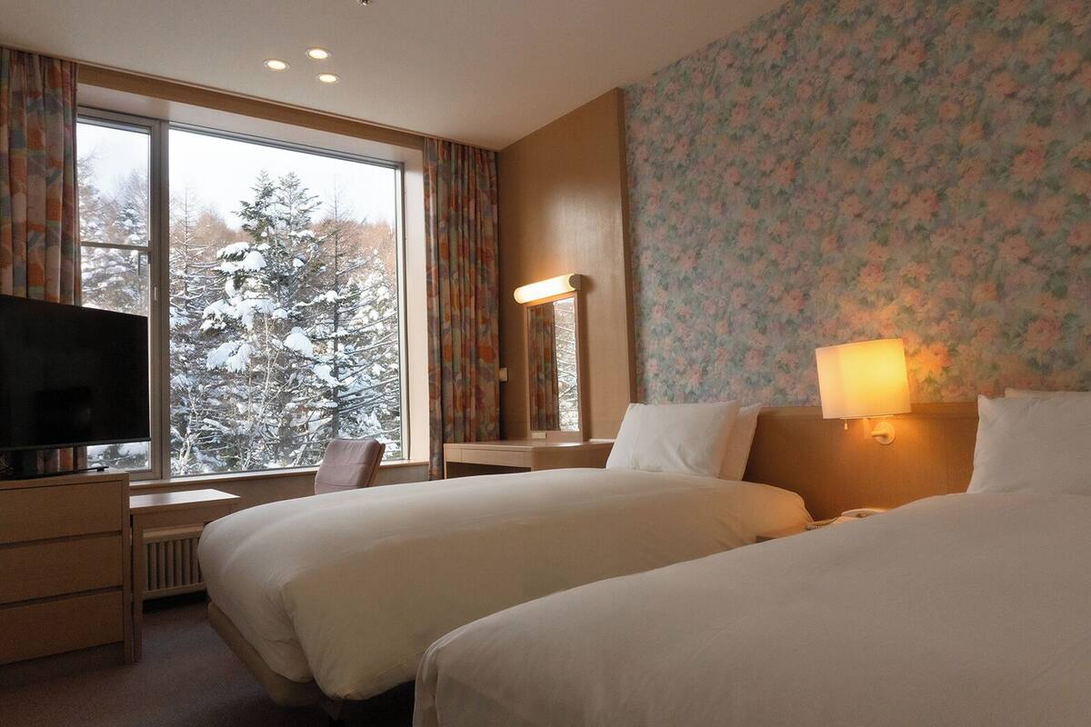 Habitación de hotel con dos camas, televisión, escritorio y ventana con vistas a árboles nevados.