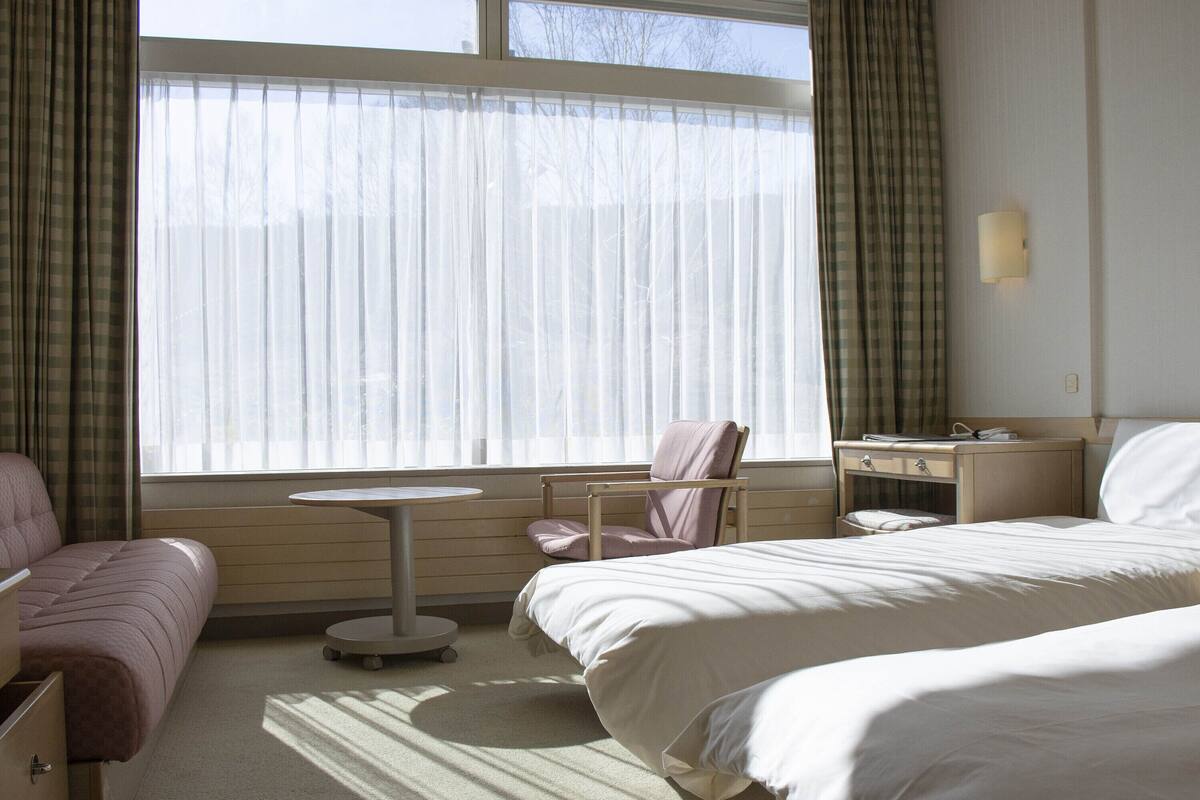 Habitación de hotel con dos camas, una mesita, una silla, una mesita de noche y un ventanal con cortinas.