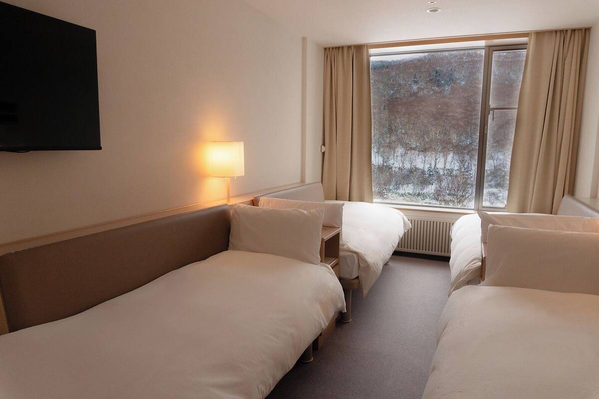Habitación de hotel con dos camas, un televisor montado en la pared, una ventana con cortinas y una lámpara en la mesita de noche.