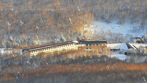 A large building with multiple windows, surrounded by snow-covered trees and a hilly landscape.