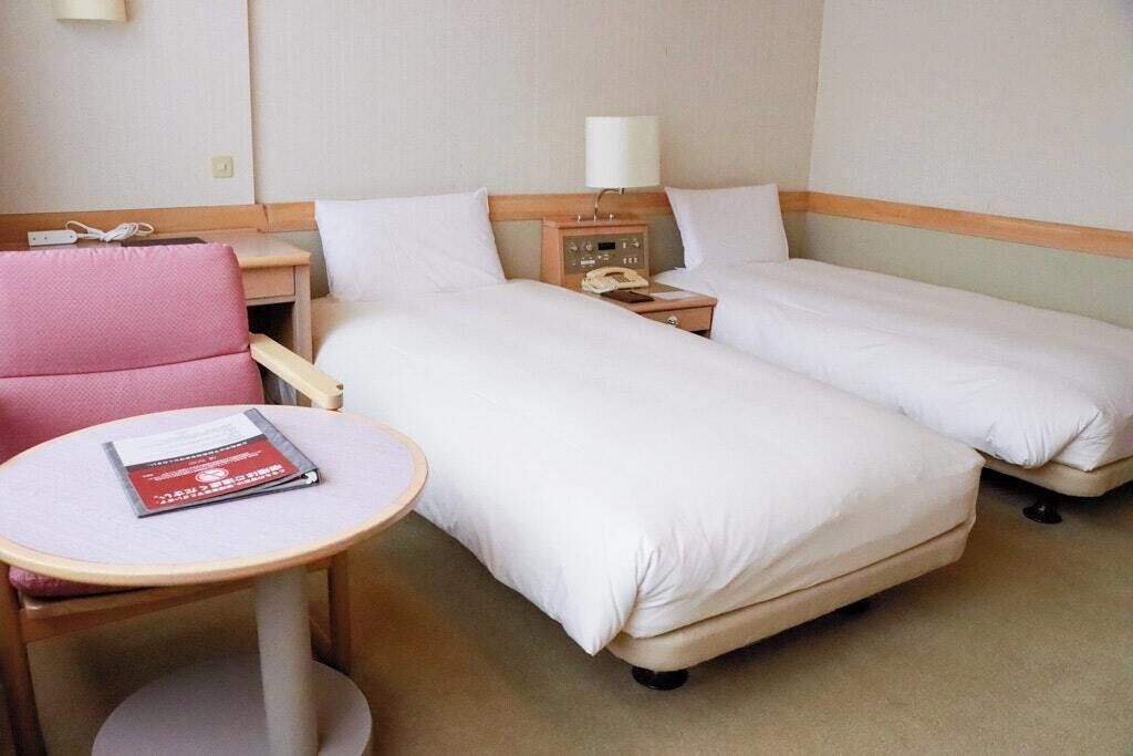Habitación de hotel con dos camas, una mesita con un libro, una silla, una mesita de noche con lámpara y un teléfono.