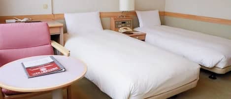 Habitación de hotel con dos camas, una mesita con un libro, una silla, una mesita de noche con lámpara y un teléfono.