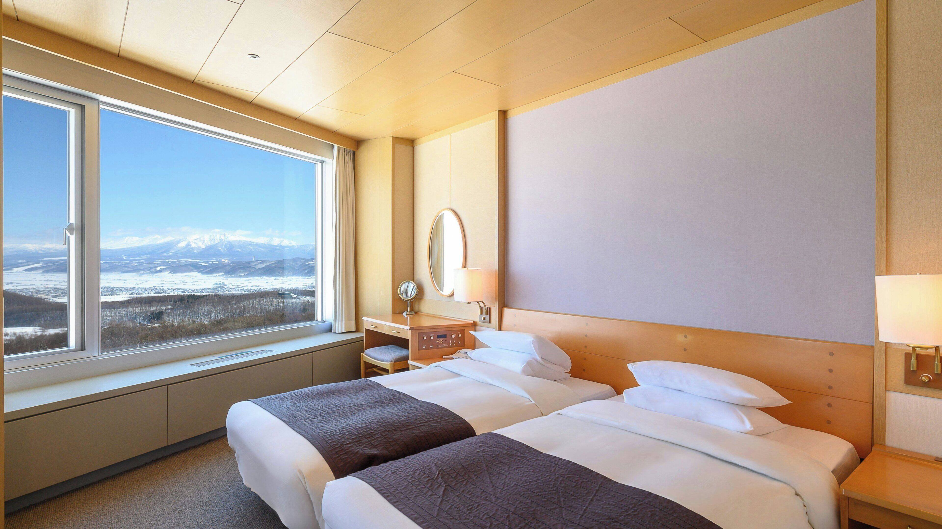 A hotel room with a large window, a bed, a bedside table, a mirror, and a lamp.