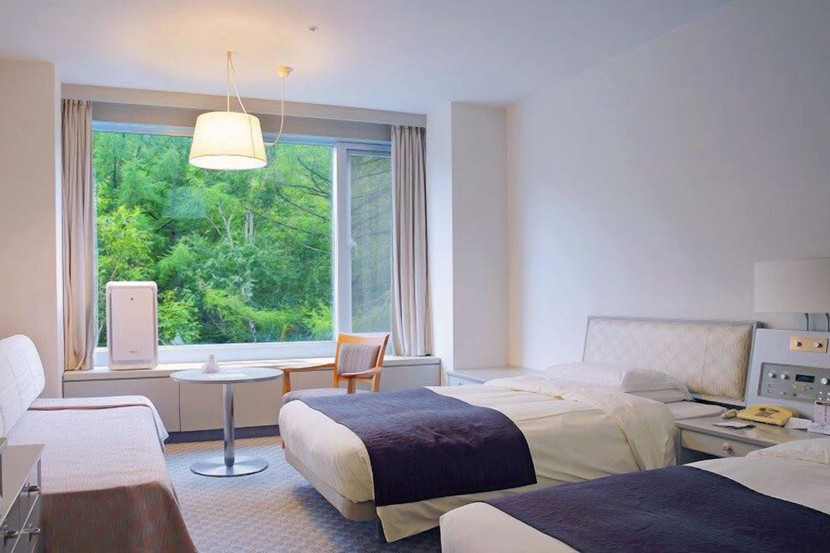 Quarto de hotel com duas camas, uma escrivaninha, uma cadeira e janela com vista para o verde.