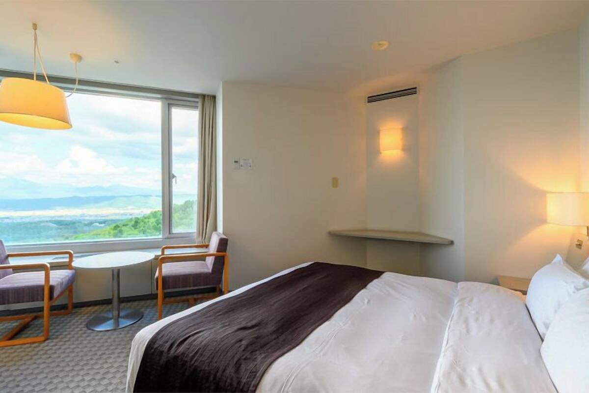 Quarto de hotel com uma cama, duas poltronas, uma mesinha e vista para o oceano.