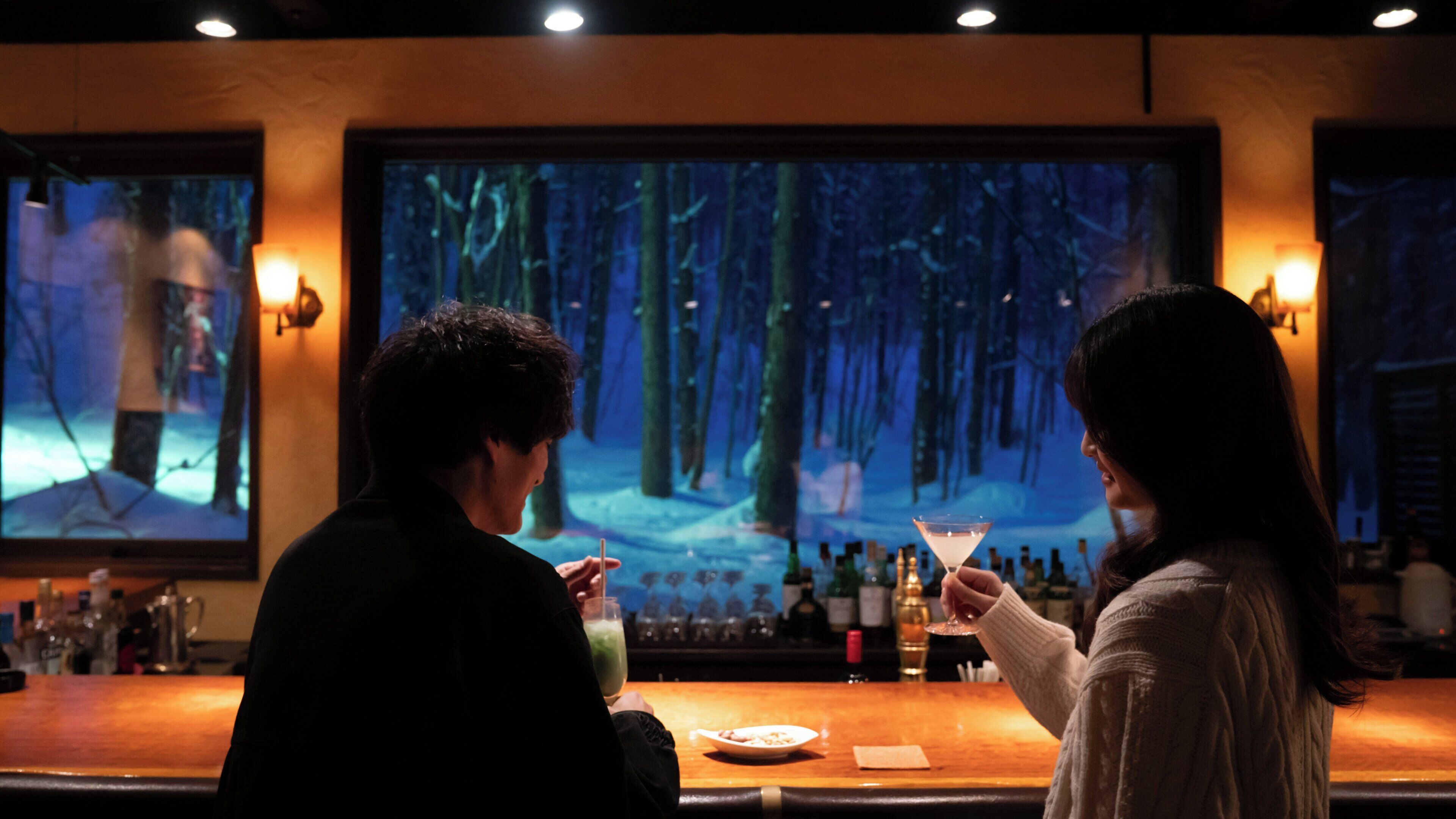 Dos personas en un bar, con una pantalla grande que muestra un paisaje forestal.