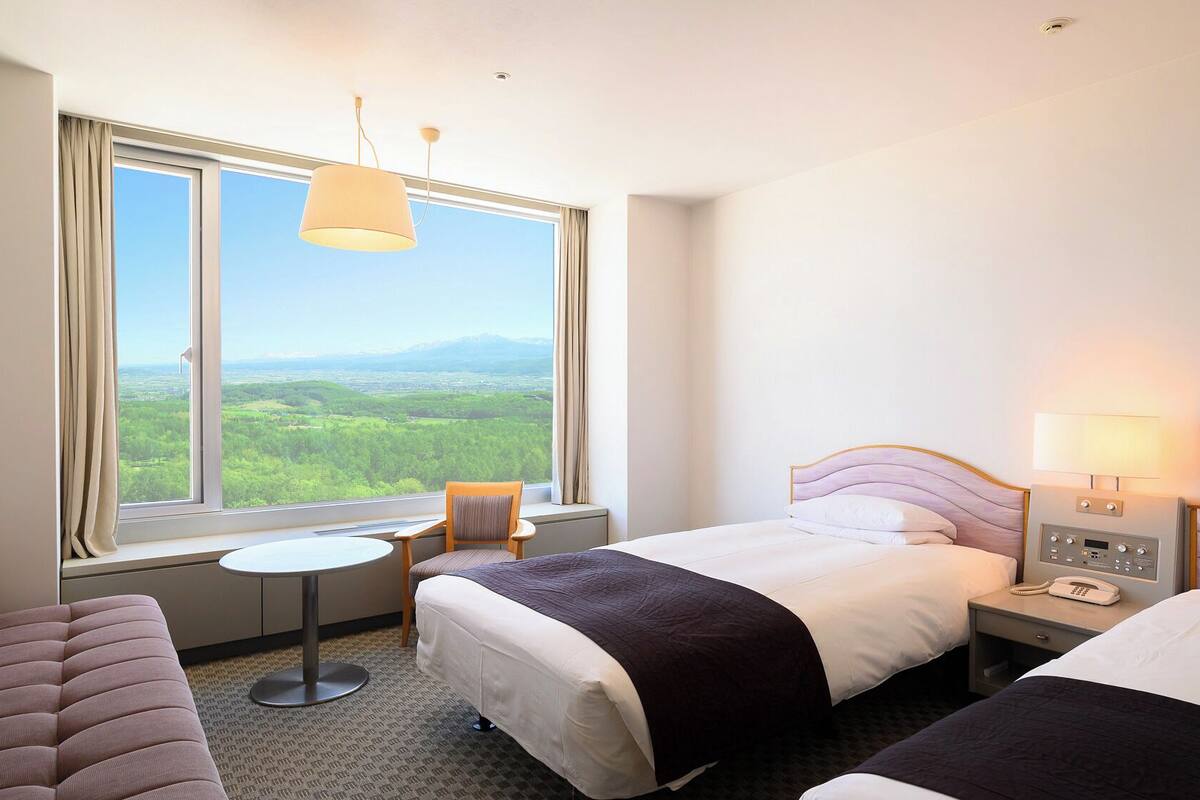 Quarto de hotel com duas camas, uma mesa pequena, uma cadeira e vista para uma paisagem verde através de janelas amplas.