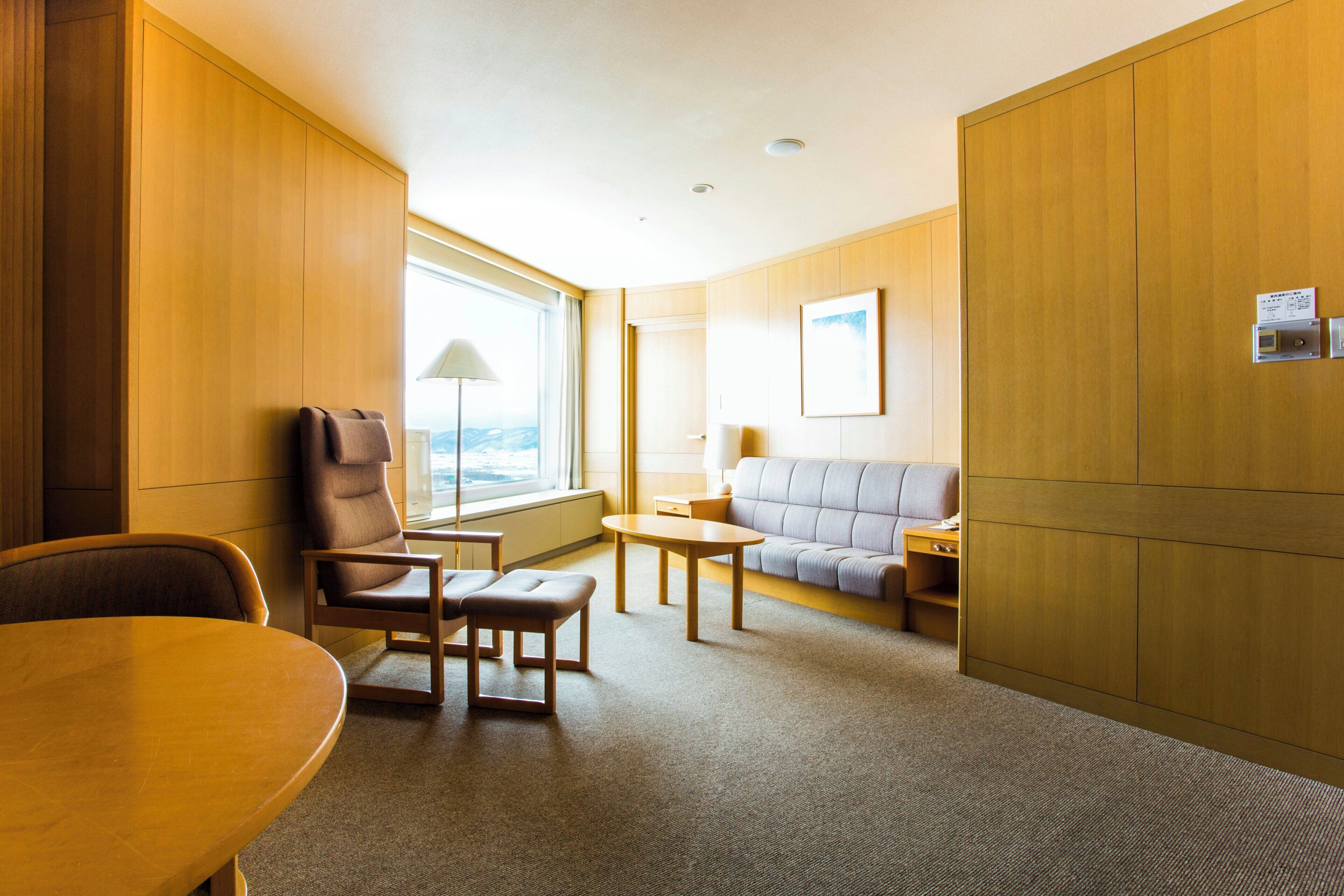 A hotel room with a wooden wardrobe, a sofa, a small table, a chair, and a desk.