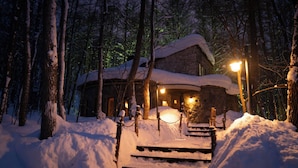 Una cabaña cubierta de nieve con fachada de piedra, escalones de madera que conducen a la entrada y una luz iluminando el frente.