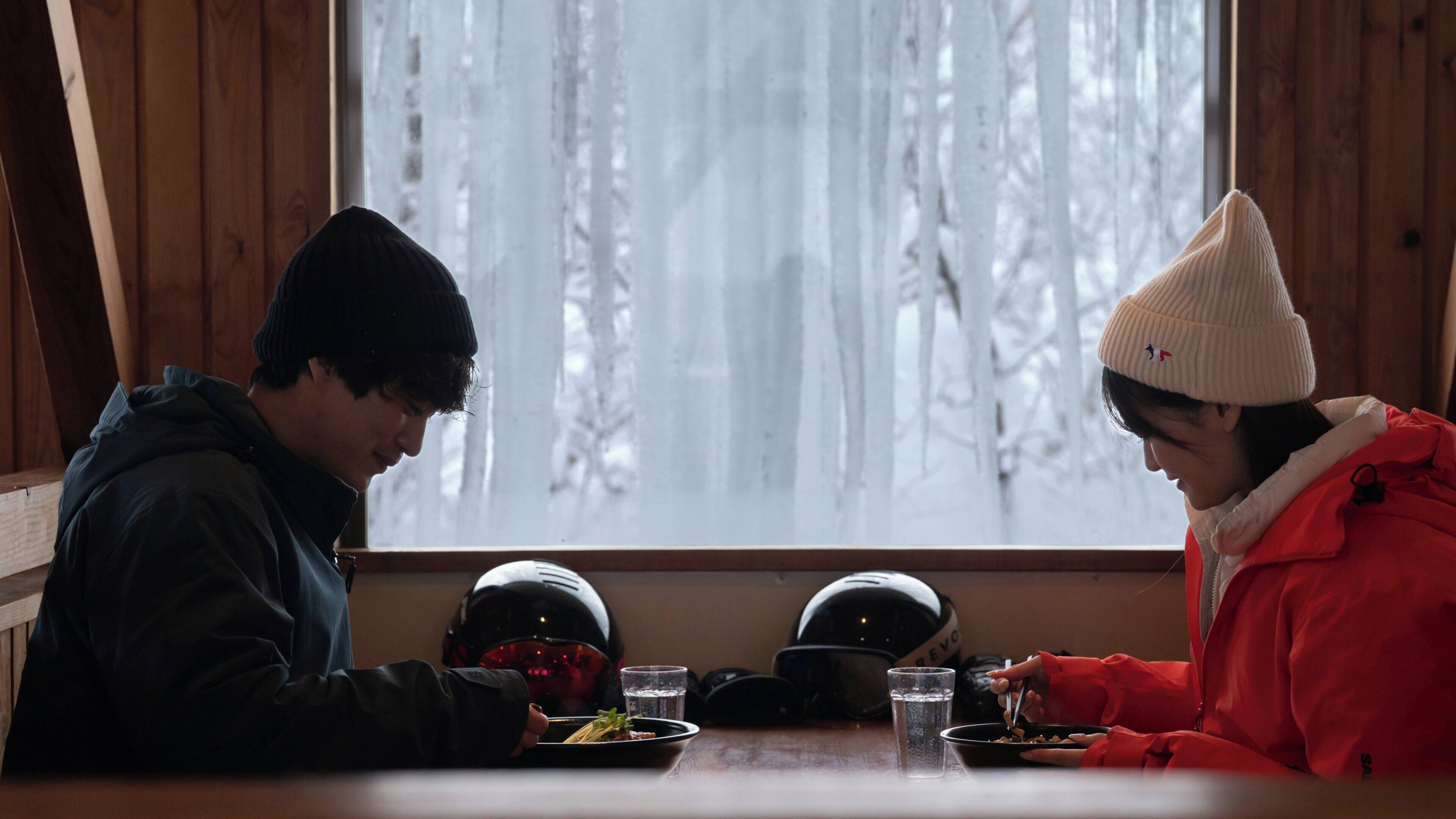 Dos personas sentadas en una mesa con cascos y un bol de comida, en una habitación con paredes de madera y una ventana con vistas a árboles.