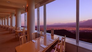 Comedor con mesas y sillas, ventanas amplias con vistas a una ciudad y montañas.