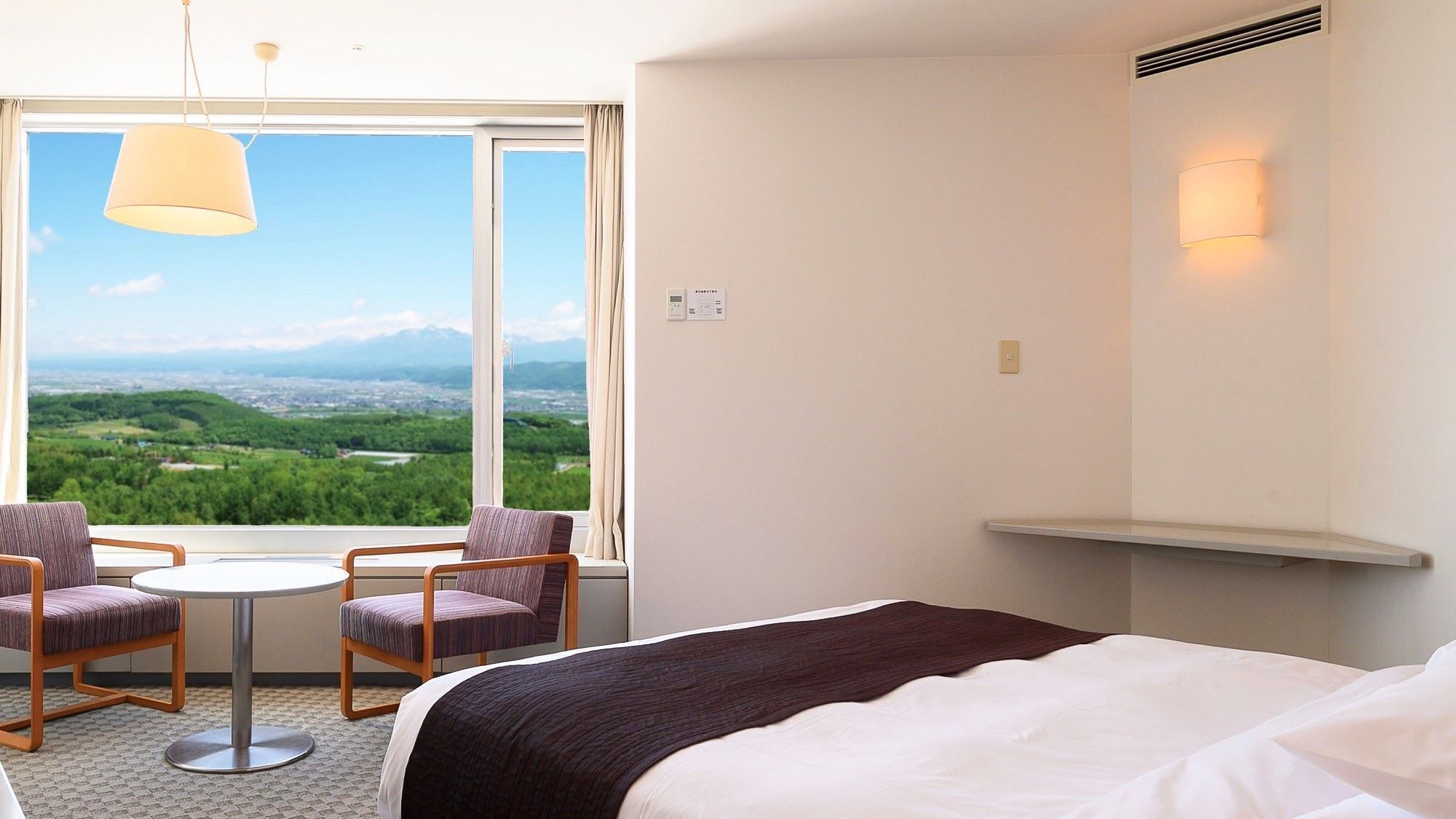 A hotel room with a bed, two chairs, a small table, a view of a green landscape, and a window with a curtain.