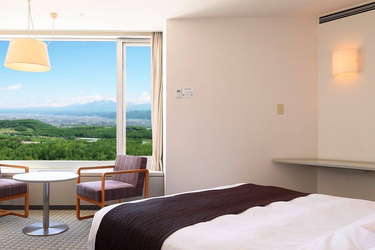 Quarto de hotel com cama, duas cadeiras, uma mesinha, vista para uma paisagem verde e uma janela com cortina.
