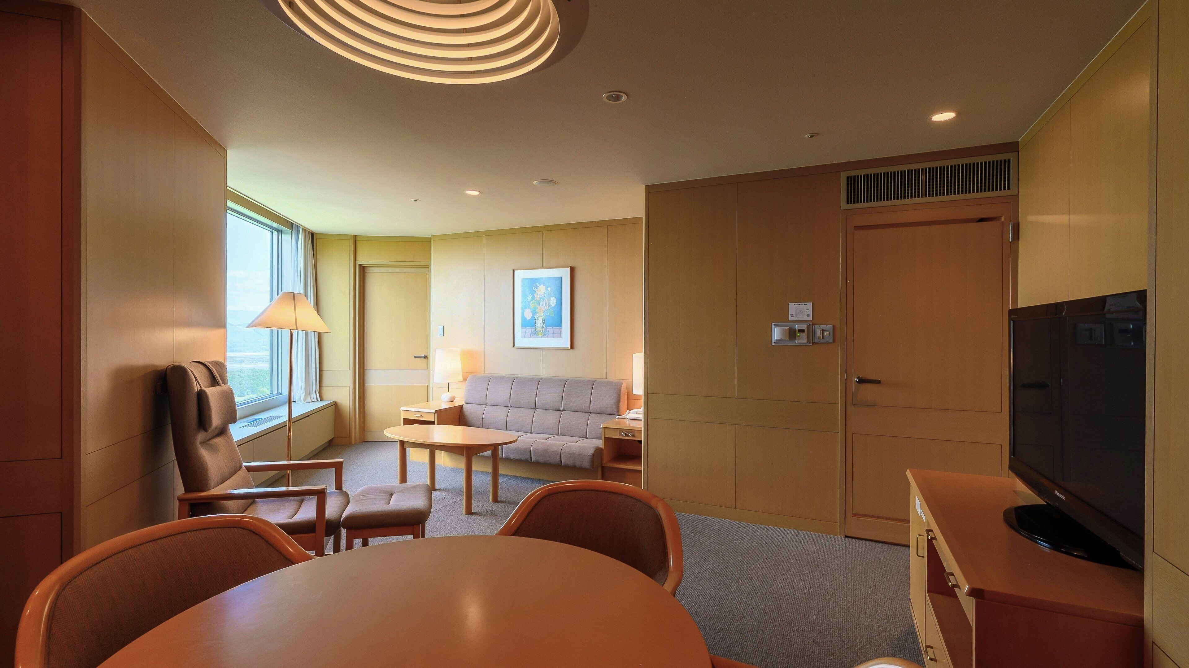 Ein Hotelzimmer mit einer Couch, einem Esstisch, einem Fernseher und einem Fenster mit Vorhängen.