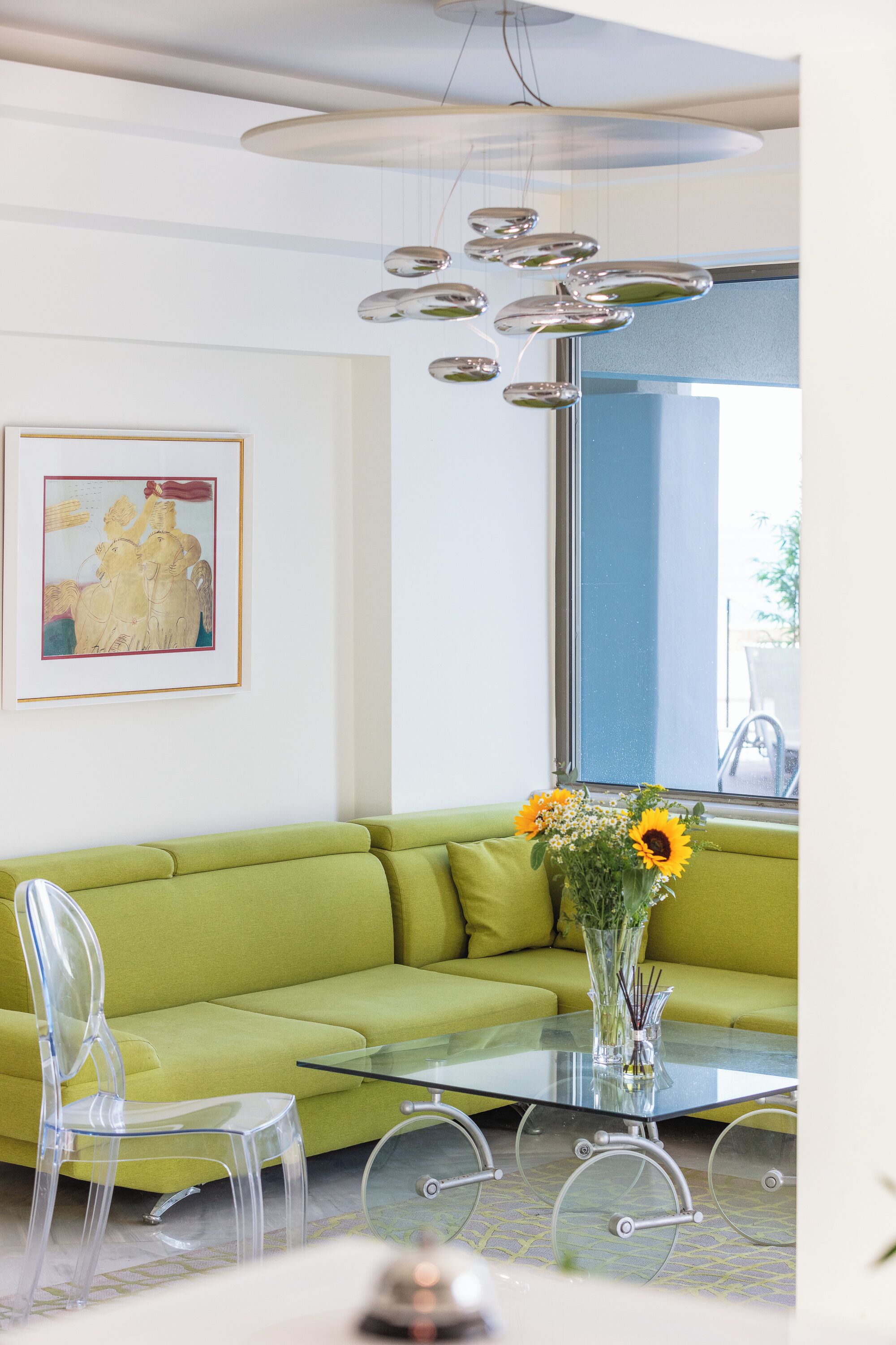 A modern living room with a green sofa, clear plastic chairs, a glass coffee table, and a vase with flowers.