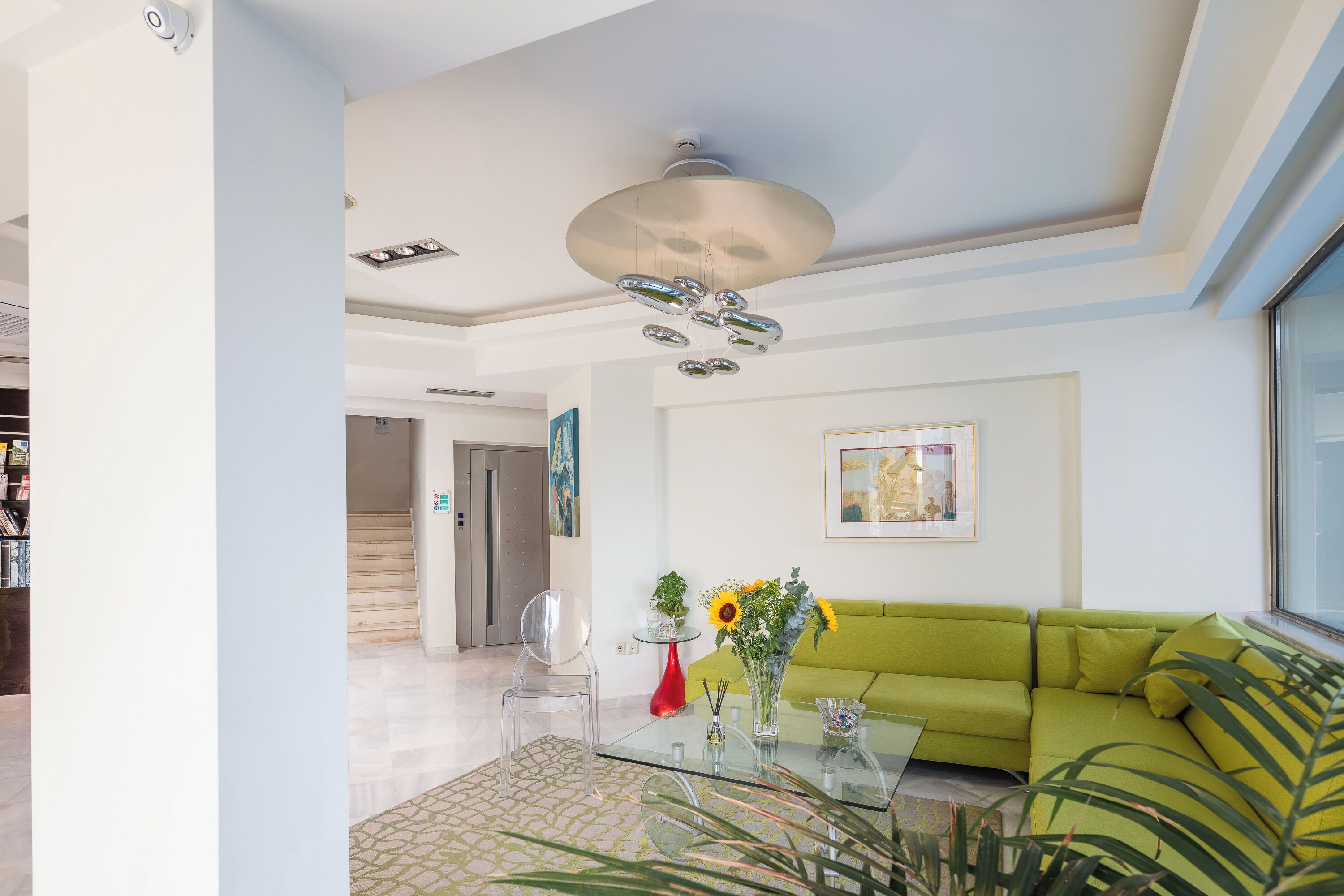 Uma sala de estar moderna com um sofá verde, uma mesa de centro de vidro e uma cadeira transparente. Há um vaso vermelho com flores em uma mesinha de cabeceira, e um lustre no teto.
