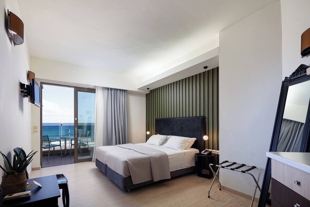 Une chambre d’hôtel moderne équipée d’un lit, d’un bureau, d’une chaise, d’une petite table, d’une télévision et offrant une vue sur la mer grâce à une porte vitrée.
