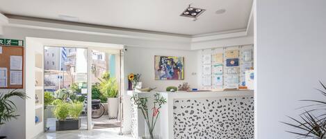 A reception area with a glass door leading outside, a reception desk with a patterned backdrop, and potted plants.