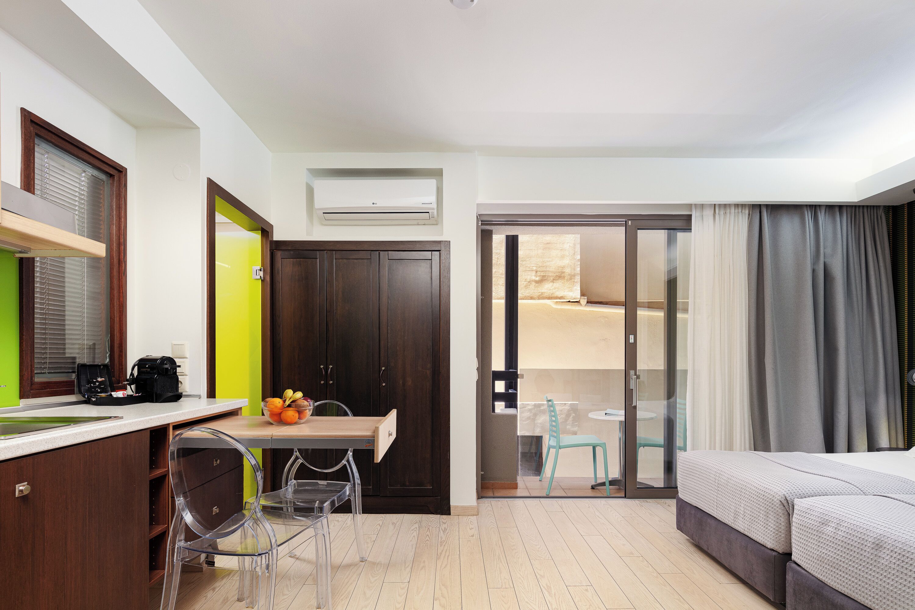 A hotel room with a bed, a desk with a chair, a small table with fruit, a wardrobe, and a sliding glass door to a balcony.