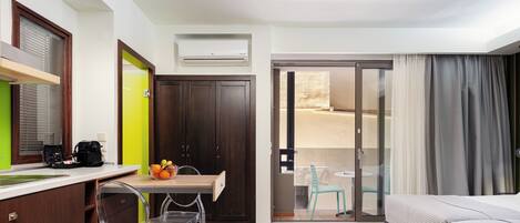 A hotel room with a bed, a desk with a chair, a small table with fruit, a wardrobe, and a sliding glass door to a balcony.