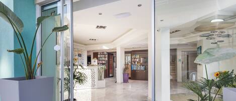 A modern hotel lobby with a reception desk, a large potted plant, and a clear glass door leading to the interior.