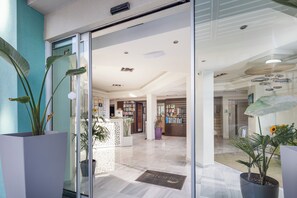 Een moderne hotellobby met een receptiebalie, een grote potplant en een glazen deur die naar de binnenkant leidt.