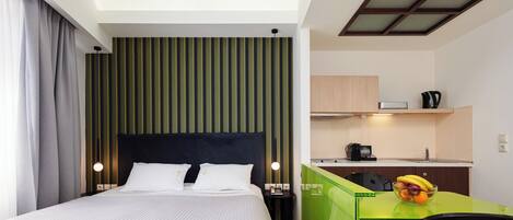 A hotel room with a bed, a desk with a bowl of fruit, a kitchenette, and a window with curtains.